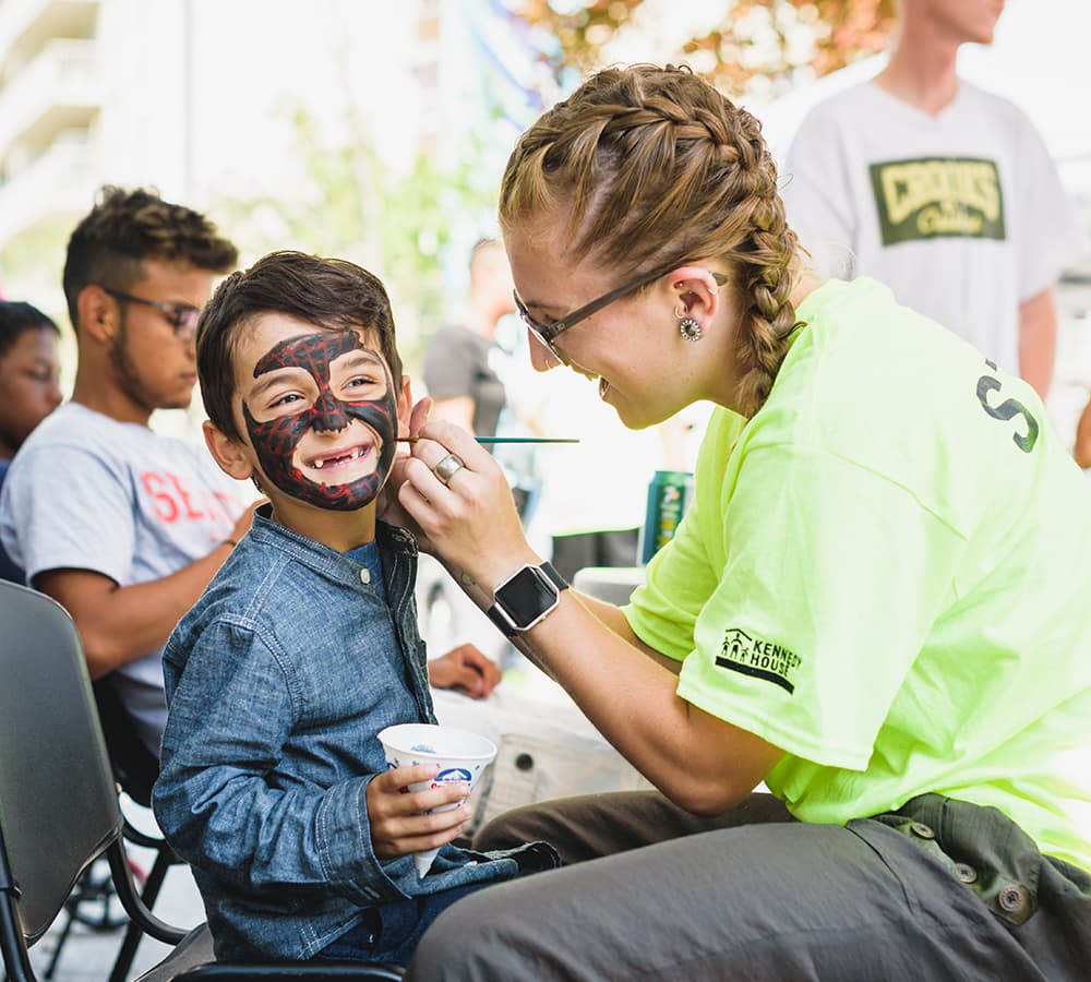 A woman in a green shirt painting a Spider Man face onto a child