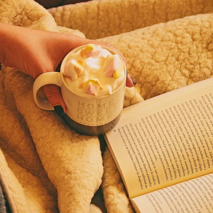 A cup of hot chocolate placed beside an open book on top of a cozy blanket